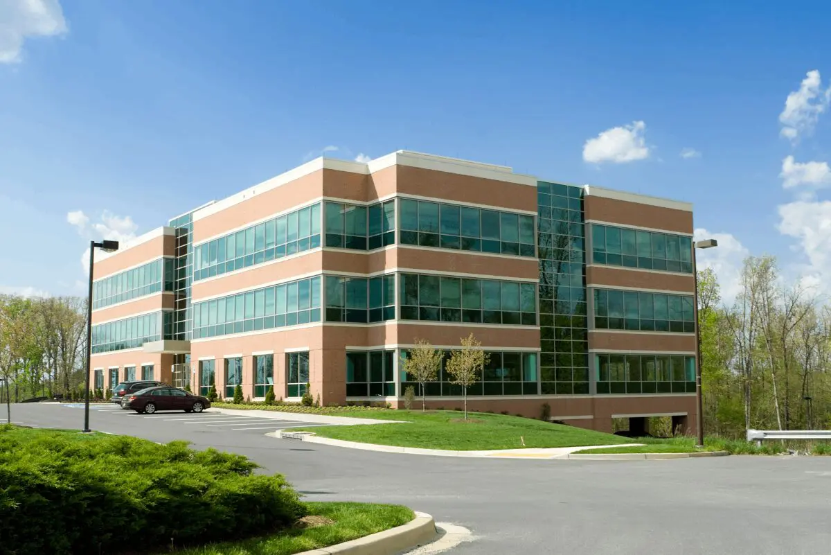 A modern, three-story office building with large windows, a brick facade, and a small parking lot with one parked car on a sunny day.