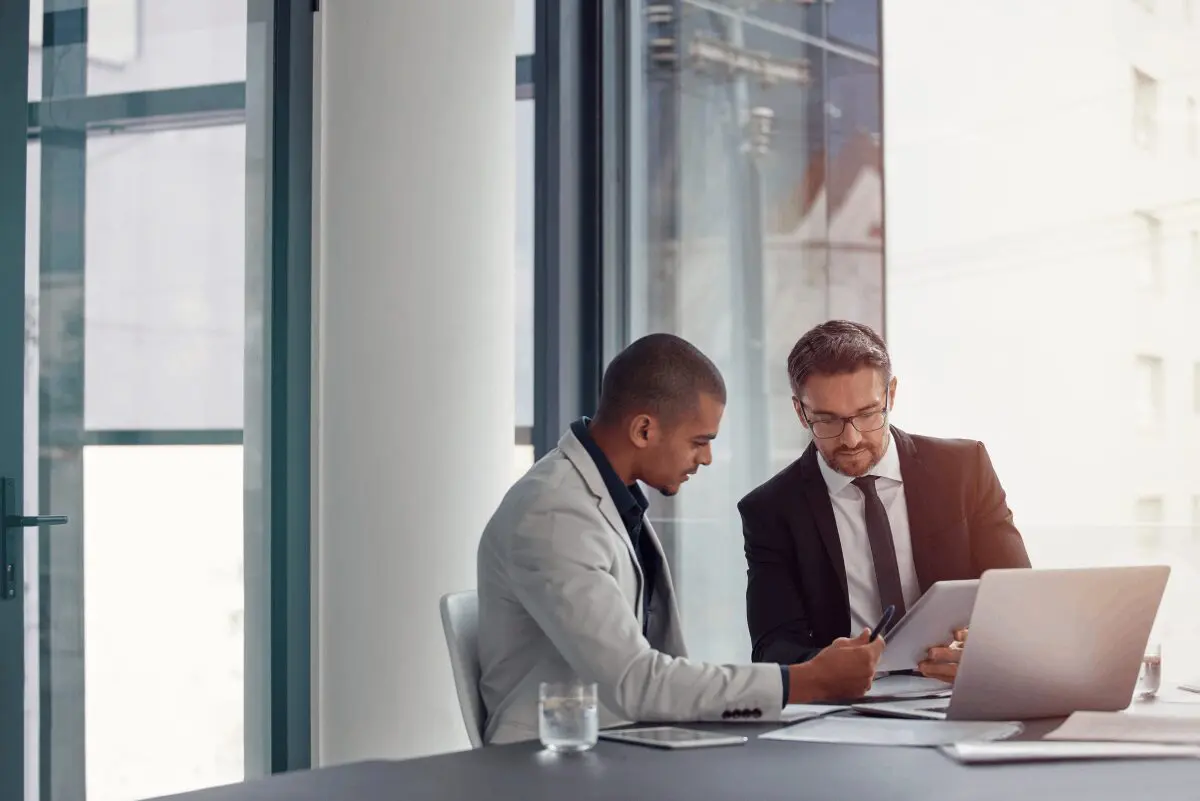 Two businessmen in suits sit at a table in a modern office, discussing documents and using a laptop and tablet.