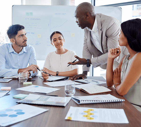Four people in business attire sit around a table with documents, notebooks, and charts, having a discussion in a modern office meeting room.