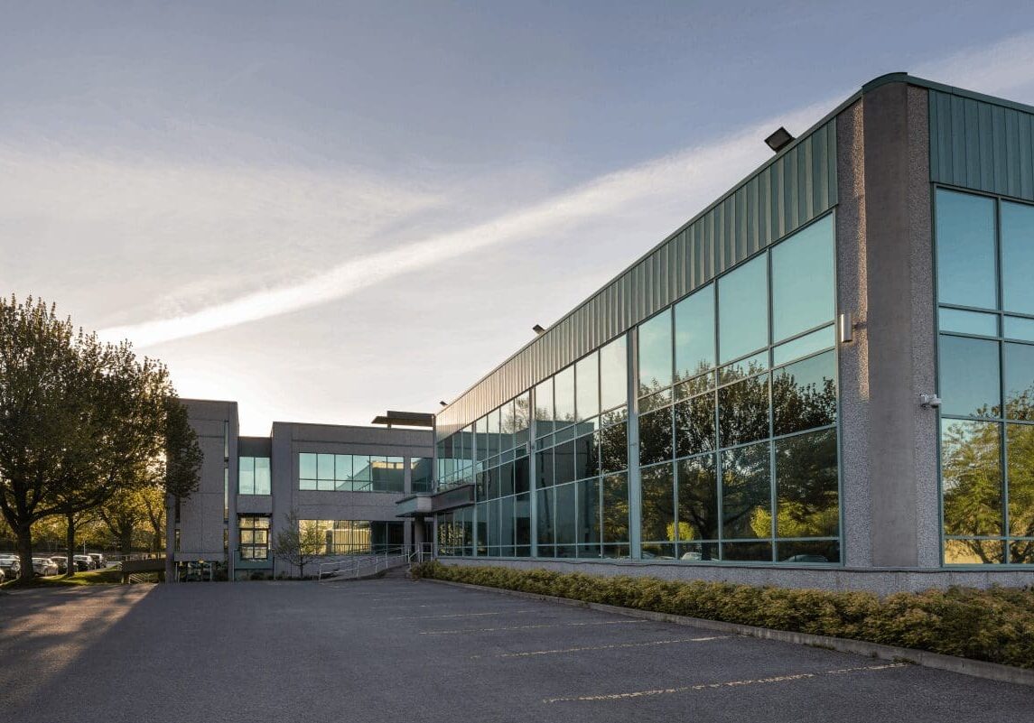 A modern, two-story office building with large reflective glass windows, surrounded by trees and an empty parking lot under a clear sky.